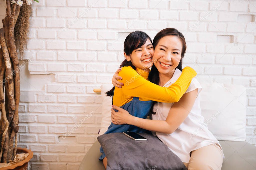 Cheerful Asian mother and daughter embracing while sitting on sofa
