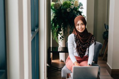 Kafede dizüstü bilgisayar kullanan gülümseyen Müslüman kadın.