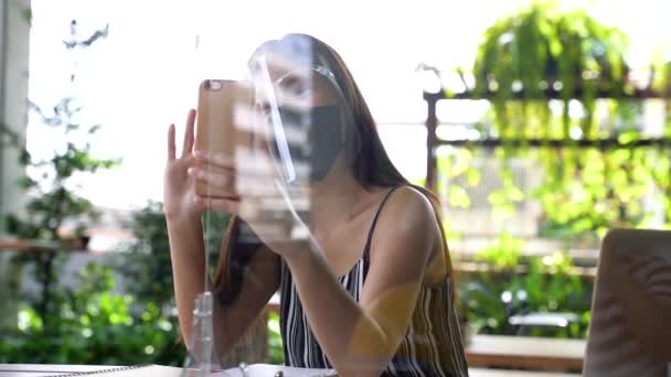 Jovem de 20 anos mulher bonita asiática vestindo um protetor facial e máscara com partição no restaurante café. Ao usar computador portátil e telefone celular fora. - Conceito de prevenção de vírus Corona — Vídeo de Stock