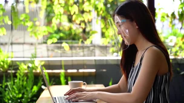아시아 여성이 노트북을 사용하며 앉아 있는 동안 얼굴 가리개를 착용하고 있습니다. 야외 사무실에서 컴퓨터를 사용 한다. 얼굴 가리개는 코로나 바이러스 - 아시아의 코 비투스 19를 예방하는 인기있는 장비이다 — 비디오