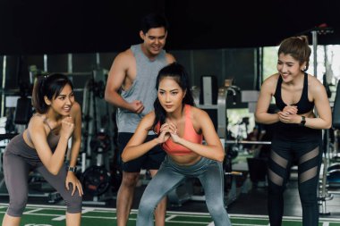 Bir grup insan, spor salonunda ağırlık plakasıyla çömelme hareketi yapan Asyalı bayan arkadaşına tezahürat yapıyor. Takım çalışması olarak çalışıyoruz..