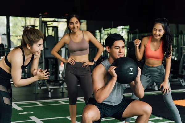 Gruppe von Menschen, die ihrem asiatisch-chinesischen Freund beim Kniebeugen mit einem Medizinball im Fitnessstudio zujubeln. Gemeinsam als Team arbeiten. — Stockfoto