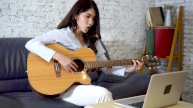 Genç Asyalı kadın dizüstü bilgisayar monitöründen gitar çalmayı öğreniyor. Kadın gitarist çevrimiçi özel ders izliyor.