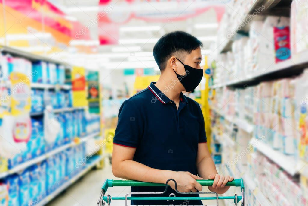 Young adult Asian man wearing a face mask while shopping with cart trolley in grocery supermarket store.