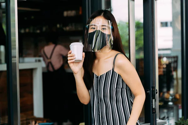 喫茶店を出発しながら外科的フェイスマスクをした若いアジア系女性。魅力的な女性の顧客はコーヒーを購入し、使い捨てのコーヒーカップを保持します。Covid-19予防 — ストック写真