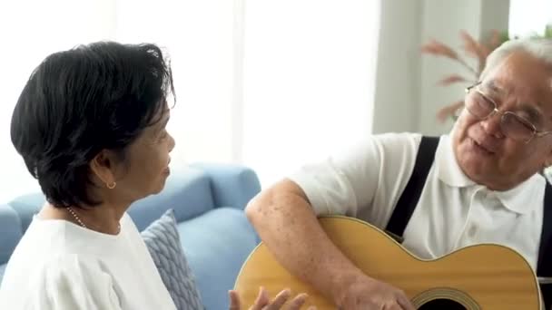 아시아인 졸업반 커플 이 소파에 앉아 음향 기타를 함께 연주하고 있습니다. 70 세의 기타리스트 남편 이 노래를 부르는 동안 나이든 여성 이 손뼉을 치며 행복하게 웃고 있습니다. 집에서 퇴직 생활을 즐김. — 비디오