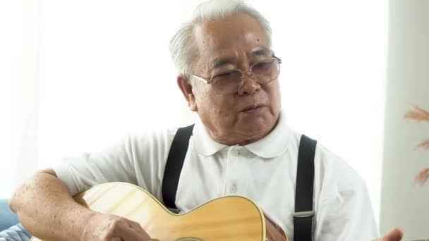 Hombre mayor asiático sentado en el sofá y tocando la guitarra acústica. Feliz anciano sonriente cantando y disfrutando con guitarra. Disfrutar de la vida de jubilación en casa. — Vídeo de stock