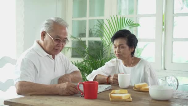 Pareja asiática mayor desayunando juntos en el comedor. 70 jubilado anciano hombre y mujer leyendo periódico — Vídeo de stock