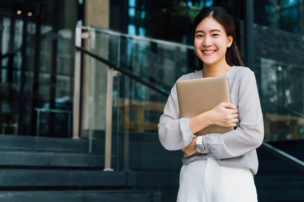 One successful and happy young adult business woman holding a laptop with smile. Professional female worker standing in corporate modern office and window in casual gray look. - With copy space — Stock Photo, Image