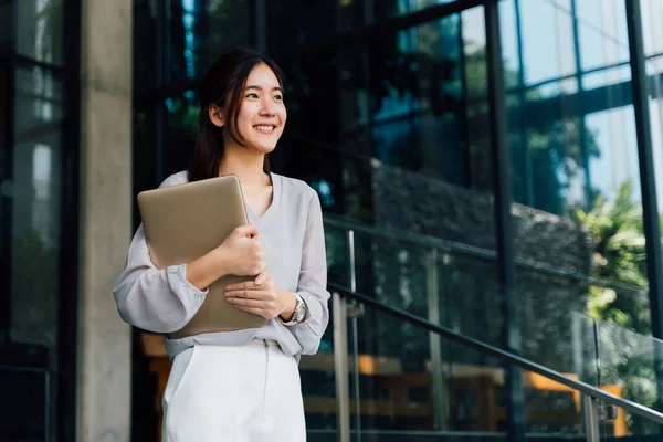 One successful and happy young adult business woman holding a laptop with smile. Professional female worker standing in corporate modern office and window in casual gray look. - With copy space — Stock Photo, Image