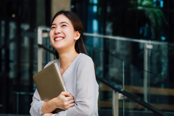 One successful and happy young adult business woman holding a laptop with smile. Professional female worker standing in corporate modern office and window in casual gray look. - With copy space — Stock Photo, Image