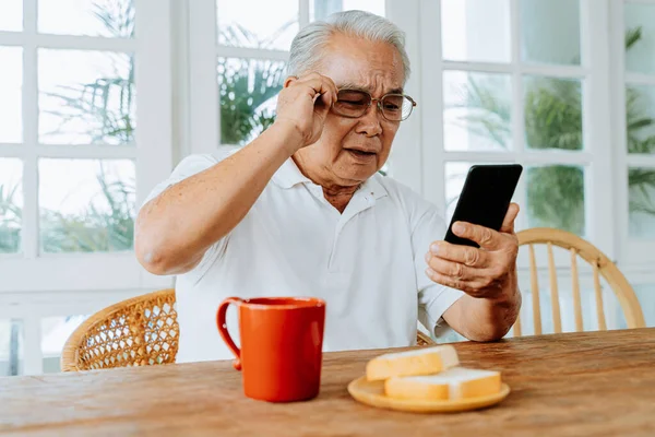 Elderly Asian male having eyesight problems and wearing eyeglasses while gazing and reading message on smartphone during breakfast at home. Old man having eye blurred vision indoors