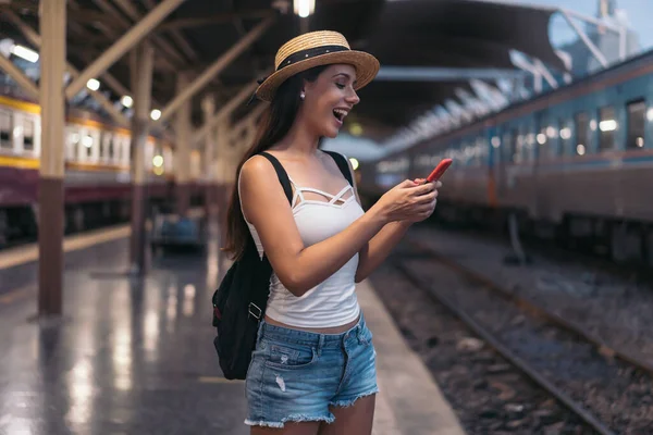 Young brunette woman traveler laughs while playing a mobile phone in train station platform. — Stock Photo, Image