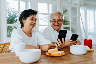 Mutlu Asyalı kadın ve erkek mutlu bir şekilde birlikte kahvaltı ederken sosyal medyaya bakıp gülümsüyorlar. Evde cep telefonu kullanan yaşlı çift.