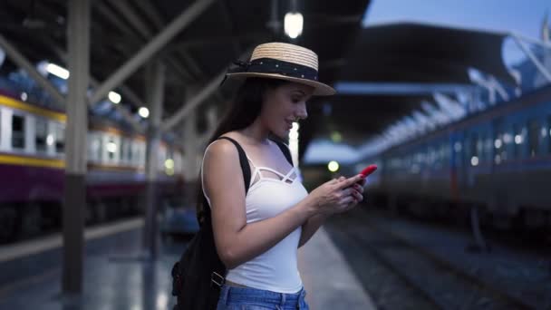 Joven morena viajero se ríe mientras juega un teléfono móvil en la plataforma de la estación de tren — Vídeo de stock