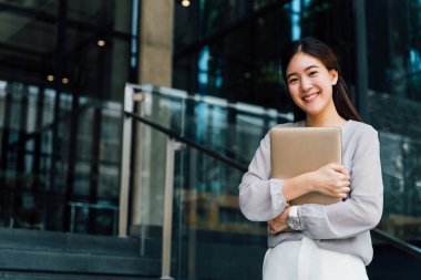 Başarılı ve mutlu bir iş kadını elinde gülümseyerek dizüstü bilgisayar tutuyor. Günümüz modern ofisinde ve penceresinde sıradan gri görünümlü profesyonel bir kadın işçi. - Kopyalama alanı ile