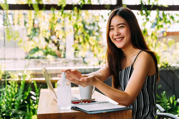 20-an muda wanita cantik Asia dengan senyum menarik menggunakan gel pembersih tangan untuk mencuci tangannya. Sambil menggunakan laptop komputer dan ponsel di luar adegan kafe. Konsep pencegahan virus Corona — Stok Foto