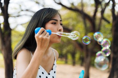 Güneydoğu Asya kökenli 20 'li yaşlardaki genç bir kadın parkta hava kabarcıkları üflüyor. Mutlu kız arka planda ağaçlar olan yaz elbisesi giyer..