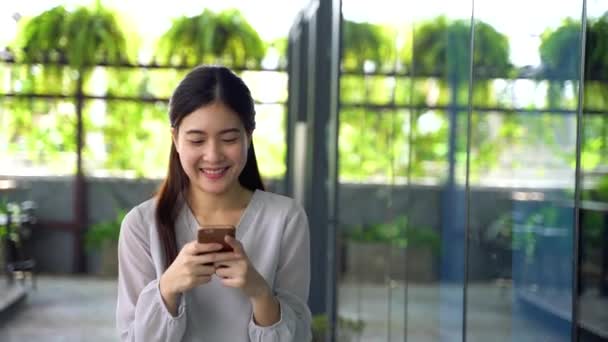 20多岁的亚洲美女在窗玻璃大楼里散步，拿着手机. — 图库视频影像
