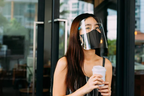 Jovem bela mulher asiática vestindo uma máscara cirúrgica ao sair de uma cafetaria. Cliente feminino atraente comprando café e segurando uma xícara de café descartável. Prevenção de Covid-19 — Fotografia de Stock