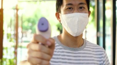 Yüz maskeli genç Asyalı adam vücut ısısını ölçmek ve izlemek için tıbbi dijital kızılötesi termometre kullanıyor. Coronavirus COVID-19 'un yayılmasını önlemek için ziyaretçileri görüntülemek üzere yeni normal yaşam