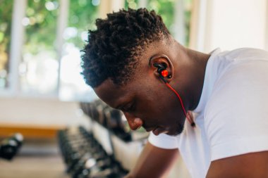 Spor kıyafetli yorgun Afrikalı Amerikalı adamın spor salonunda antrenman sırasında bankta oturup dinlenirken kablosuz kulaklıkla müzik dinlemesi.