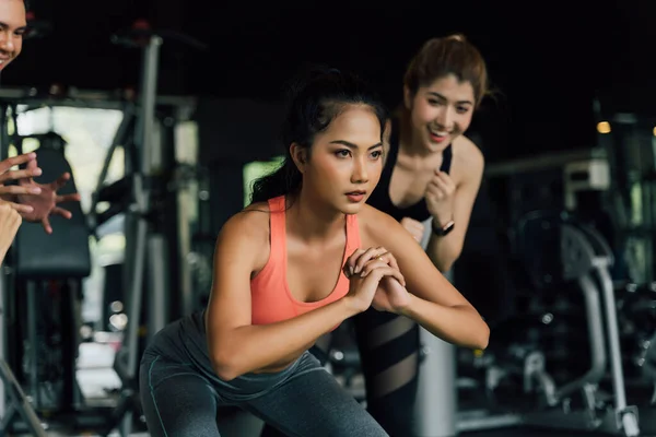 Grupo de pessoas aplaudindo sua amiga asiática fazendo agachamentos com uma placa de peso no ginásio de fitness. Trabalhando juntos como um trabalho de equipe. — Fotografia de Stock