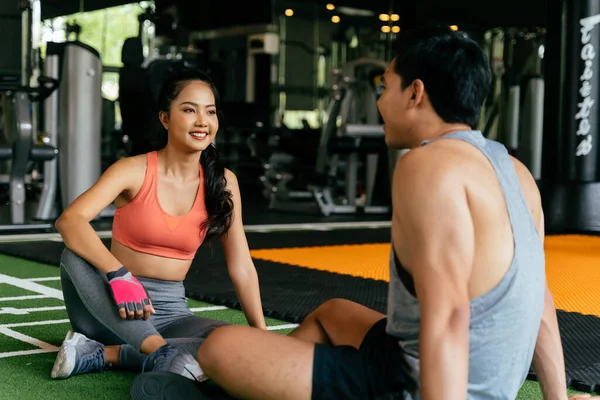 Лицо молодой азиатской смешанной спортсменки болтает с мускулистым мужчиной, сидящим на полу в фитнес-зале. — стоковое фото
