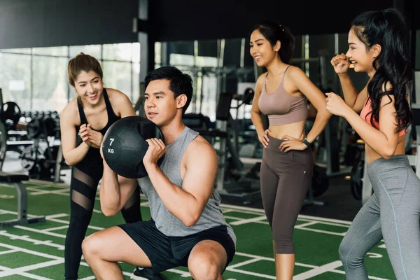 Een groep mensen juichen hun Aziatische Chinese mannelijke vriend toe die hurkt met een medicijnbal in de fitnessruimte. Samenwerken als een teamwork. — Stockfoto