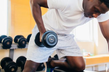 Spor kıyafetleri içinde Afrikalı Amerikalı bir adamın spor salonunda spor yaparken bençe yaslanıp, dambılla kürek egzersizi yapması.