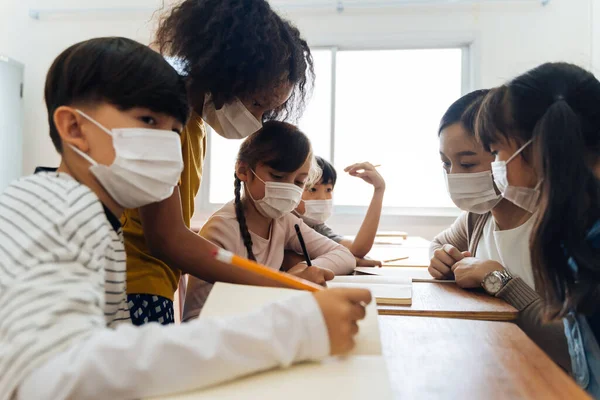 Grupa różnorodnych uczniów w budynku szkolnym omawiających na zajęciach grupowych. Podstawowe źrenice noszą maskę podczas burzy mózgów.. — Zdjęcie stockowe