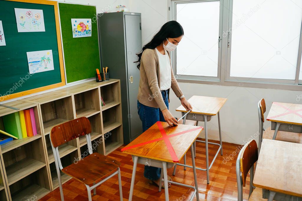 Asian female teacher wearing a face mask measures distances between desks in classroom in primary school.