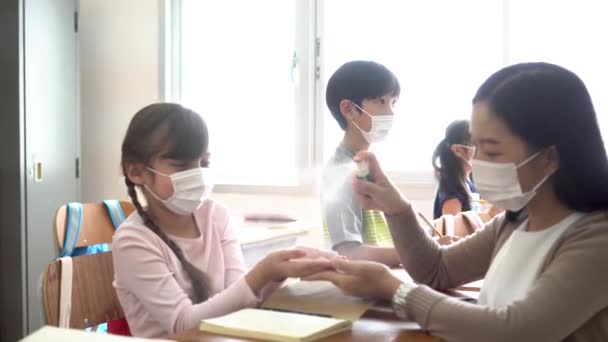 Egy fiatal tanárnő alkoholos spray-vel fertőtleníti a diákok kezét az osztályteremben. Ázsiai nő arcmaszkban kézfertőtlenítővel tisztítja a pupillákat. A karantén és a lezárás után újra megnyitják az iskolát. — Stock videók