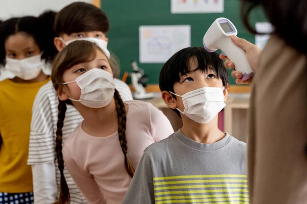 Grupa różnorodnych uczniów w budynku szkolnym sprawdziła i zeskanowała pod kątem kontroli temperatury. Uczniowie szkoły podstawowej noszą maskę i ustawiają się w kolejce przed wejściem do klasy.. — Zdjęcie stockowe