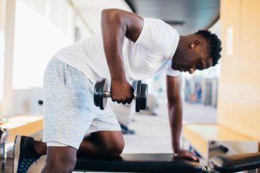 Spor kıyafetleri içinde Afrikalı Amerikalı bir adamın spor salonunda spor yaparken bençe yaslanıp, dambılla kürek egzersizi yapması.