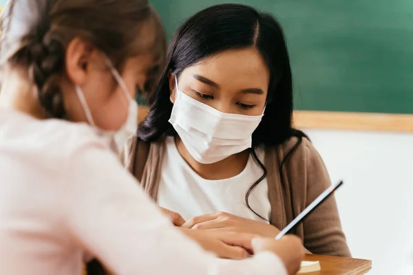 Zbliżenie azjatyckiej nauczycielki noszącej maskę na twarzy w budynku szkolnym udzielającej korepetycji uczennicy. Podstawowy uczeń pisze i uczy się w klasie.. — Zdjęcie stockowe