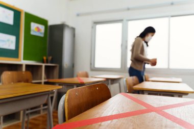 Covid19 'un yeni normal kavramı için eğitim binasında sosyal uzaklık ölçüm politikasını korumak için ilkokulda sınıfın masalarının üzerinde kırmızı bant kullanan bir bayan öğretmen.