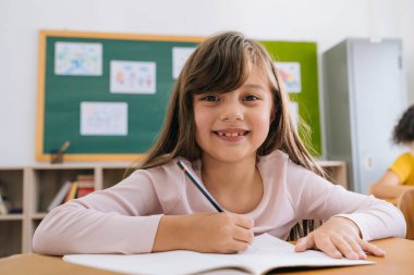 Sınıfta oturmuş gülümseyen mutlu bir kız öğrencinin portresi. Sevimli beyaz bir ilkokul öğrencisi okulda oturmuş kağıda yazı yazıyor, kameraya, eğitime, öğrenmeye, mutluluğa bakıyor.