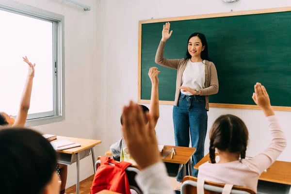 Asian school teacher with students raising hands. Young woman working in school with arm raised, school children putting their hands up to answer question, enthusiasm, eager, enjoyment — Stock Photo, Image