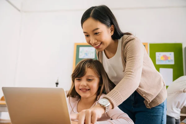 Joven maestra asiática viendo a una niña de escuela caucásica usando computadora en el aula de la escuela, nativo digital, tecnología, aprendizaje, orientación, educación. Instructora y estudiante sonriendo —  Fotos de Stock