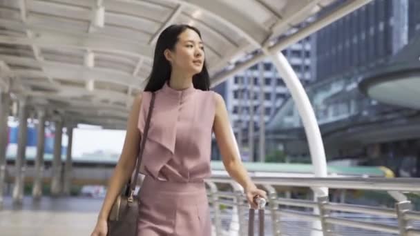 Jeune femme d'affaires asiatique marchant tout en traînant un sac trolley dans les bâtiments urbains modernes. Navetteur féminin en voyage d'affaires. — Video