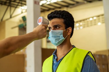 Yüz maskesi takan genç Hintli fabrika işçisi çalışmadan önce vücut ısısını kızılötesi termometre tarayıcısıyla kontrol ettiriyor. Coronavirus Covid-19 salgınında iş yerinde güvenlik önlemi