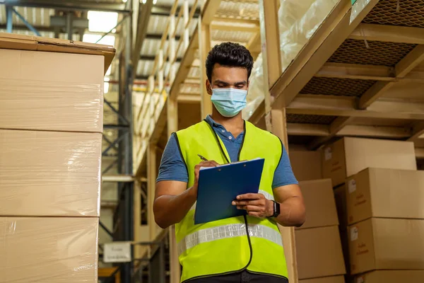 Young Indian factory warehouse worker wearing a protective face mask while working in logistic industry. 30s man checking item stock order during Covid 19 pandemic outbreak — Stock Photo, Image