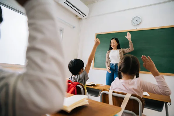 Asian school teacher with students raising hands. Young woman working in school with arm raised, school children putting their hands up to answer question, enthusiasm, eager, enjoyment