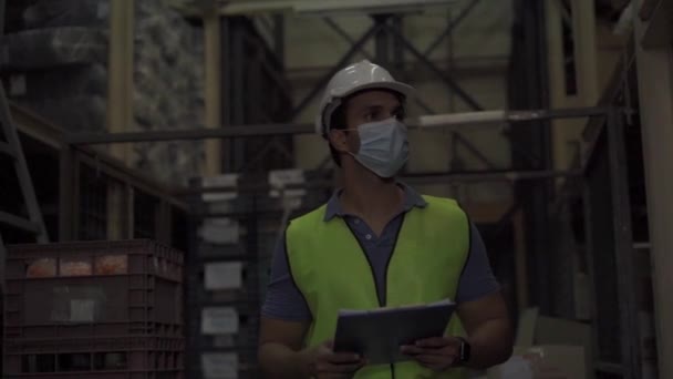 物流業界の屋内で作業しながら、保護面マスクと安全ヘルメットを身に着けている若いインドの工場倉庫労働者。コロナウイルスCovid 19パンデミックの間に30人の男が株式注文をチェック — ストック動画