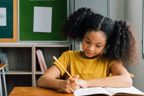 非洲裔美国女学生坐在学校里，用铅笔、学习、教育和学习来写笔记本。考试中坐在课桌前的女生 — 图库照片