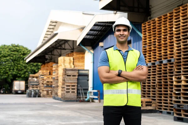 Portrait of young Indian worker with arm folded working in logistic industry in front of factory warehouse. Smiling happy man in hard hat looking at camera arms crossed at depot — Stock Photo, Image