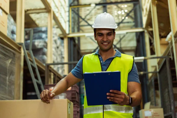 Young Indian industrial factory warehouse worker working in logistic industry indoor. Smiling happy man holding a clipboard checking merchandise stock order in storehouse