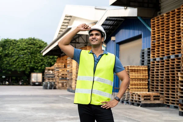 Portrait of young Indian worker working in logistic industry outdoor in front of factory warehouse. Smiling happy man with hard hat looking far away hands on hip at depot