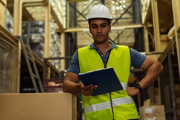 Young Indian industrial factory warehouse worker working in logistic industry indoor. Thoughtful serious man holding a clipboard checking merchandise stock order in storehouse — Stock Photo, Image